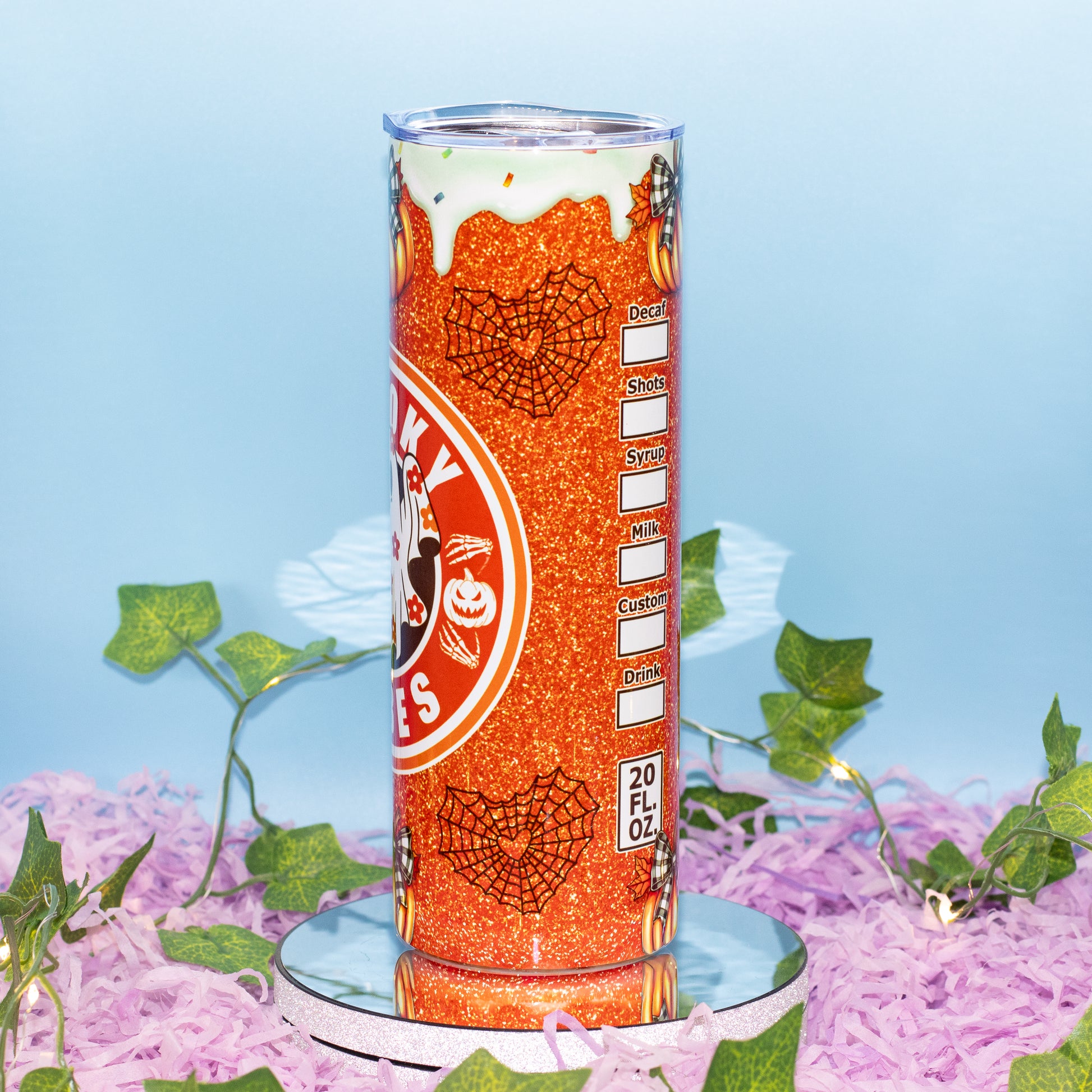 A 20 fl oz clear tumbler with a Halloween-themed orange glitter wrap, featuring spiderweb graphics, a pumpkin logo, and a white dripping frosting design near the top. The tumbler is displayed on a small mirrored pedestal surrounded by purple shredded paper and green ivy against a light blue background.