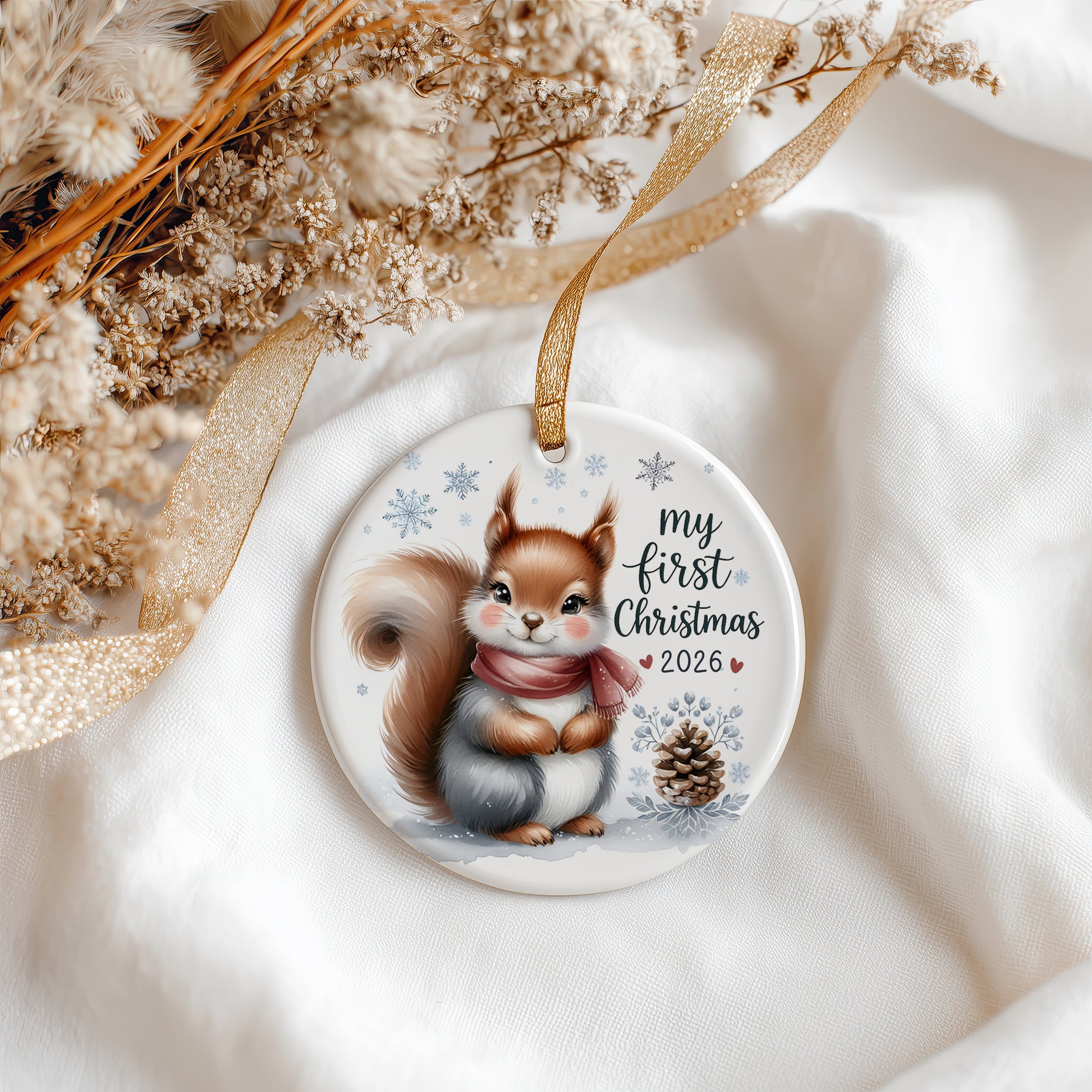 A round ceramic Christmas ornament featuring an illustration of a squirrel wearing a pink scarf, with the text 'my first Christmas 2026' and snowflakes. The ornament hangs from a gold ribbon on a soft white fabric background with dried flowers and a gold ribbon.