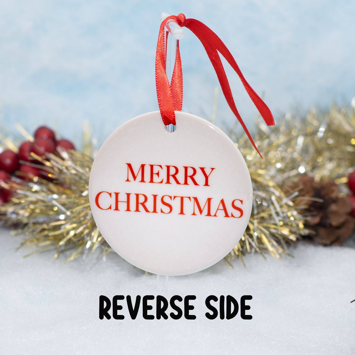 A round white Christmas ornament with red ribbon hanger, showing the front side with 'MERRY CHRISTMAS' in red text, with the caption 'REVERSE SIDE' indicating the back design. The ornament is styled with gold tinsel, pinecones, and red berries on a snowy white surface against a soft blue background.