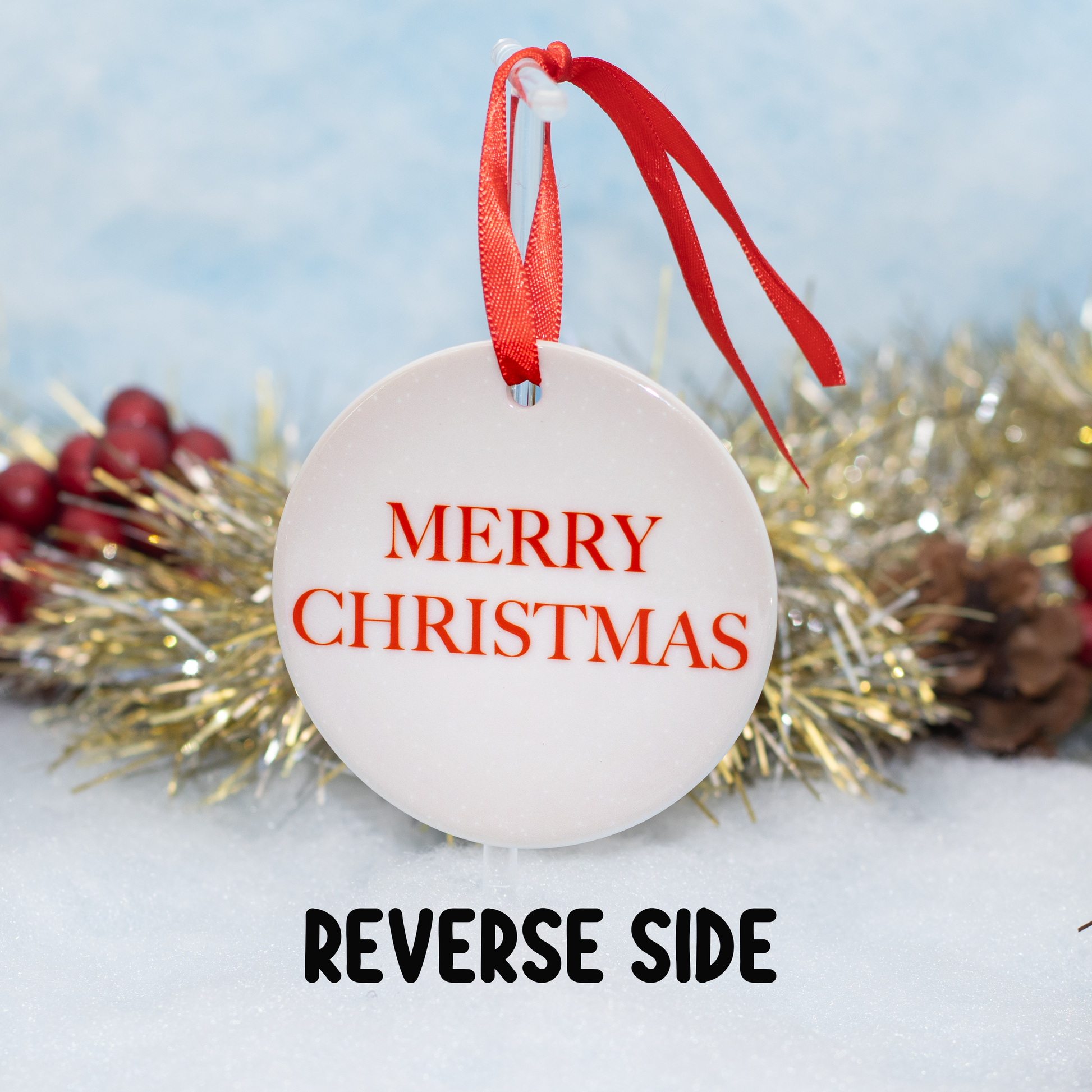 A round white Christmas ornament with red ribbon hanger, showing the front side with 'MERRY CHRISTMAS' in red text, with the caption 'REVERSE SIDE' indicating the back design. The ornament is styled with gold tinsel, pinecones, and red berries on a snowy white surface against a soft blue background.
