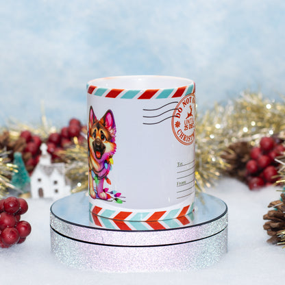 A white ceramic Christmas-themed mug featuring a colorful German Shepherd illustration and red, teal, and white postmark-style stripes, displayed on a round silver glitter pedestal with festive tinsel and red berries in the background.