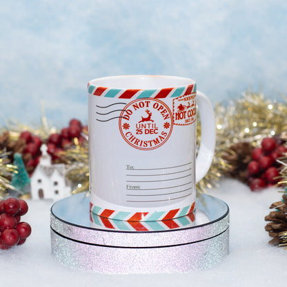 A white ceramic mug decorated with a Christmas postmark design, displayed on a round glittery pedestal with festive holiday decorations in the background.