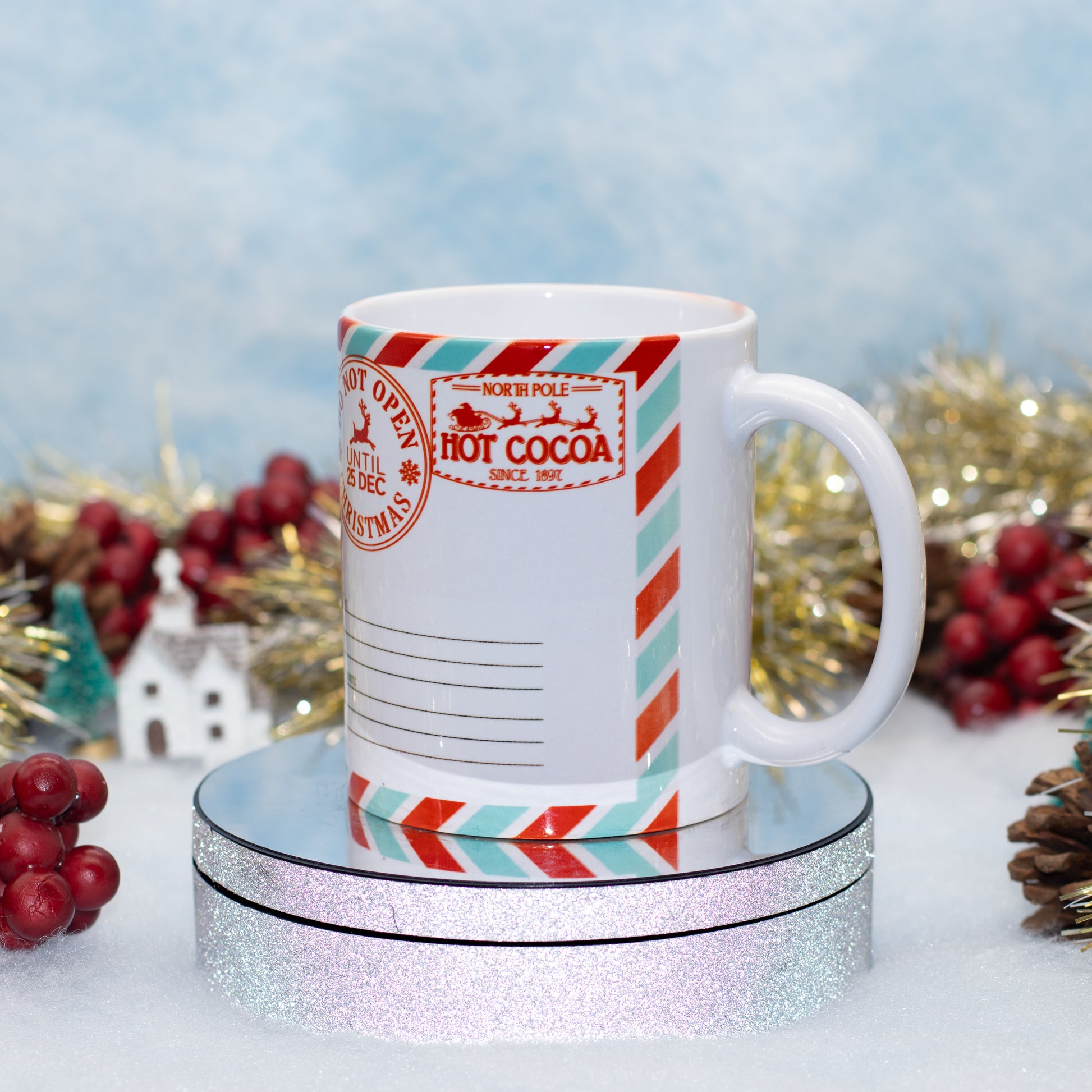 A white ceramic mug designed like a vintage North Pole hot cocoa envelope, featuring red and teal striped borders and a red stamp that reads 'HOT COCOA NORTH POLICE 1950'. The mug is displayed on a round, glittery silver pedestal with festive holiday decorations, including red berries, gold tinsel, and pinecones, against a soft, light blue textured background.