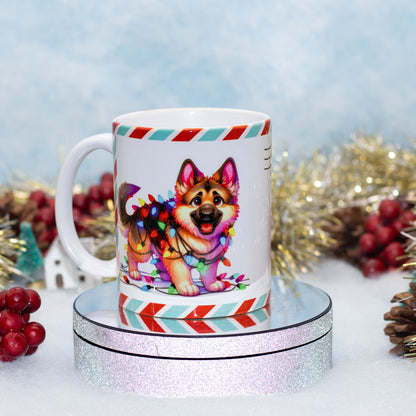 A white ceramic mug featuring a colorful illustration of a German Shepherd dog wrapped in holiday lights, displayed on a round silver glitter pedestal with festive tinsel and red berries in the background.