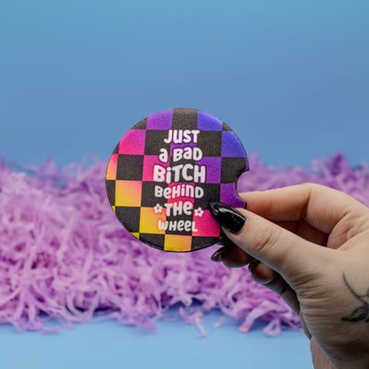 A hand holds a round car coaster featuring a colourful checkerboard pattern and the text 'JUST a BAD BITCH BEHIND THE WHEEL'. The background consists of a solid blue surface with purple shredded paper filler.