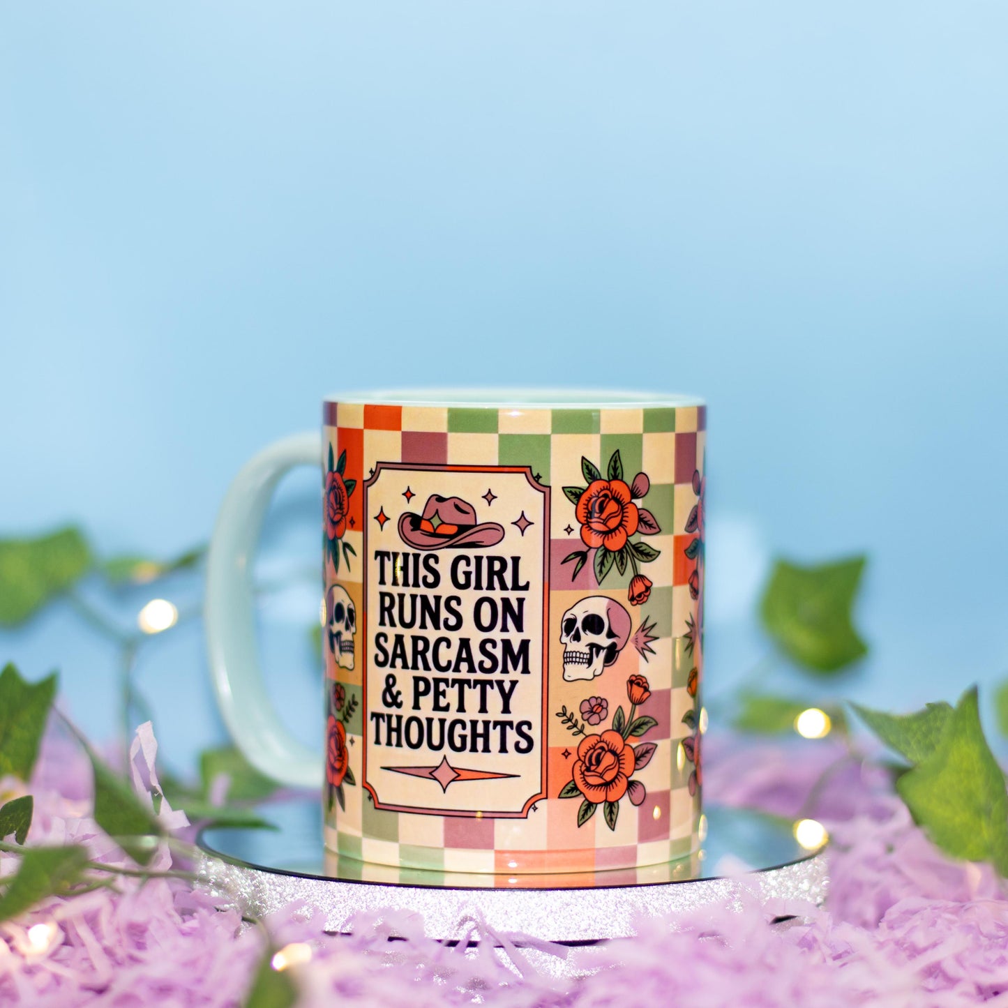 A ceramic mug with a colorful checkered border and illustrations of a cowboy hat, skull, and roses, featuring the text 'THIS GIRL RUNS ON SARCASM & PETTY THOUGHTS'. The mug is styled on a silver tray with purple shredded paper and green leaves against a soft blue background.
