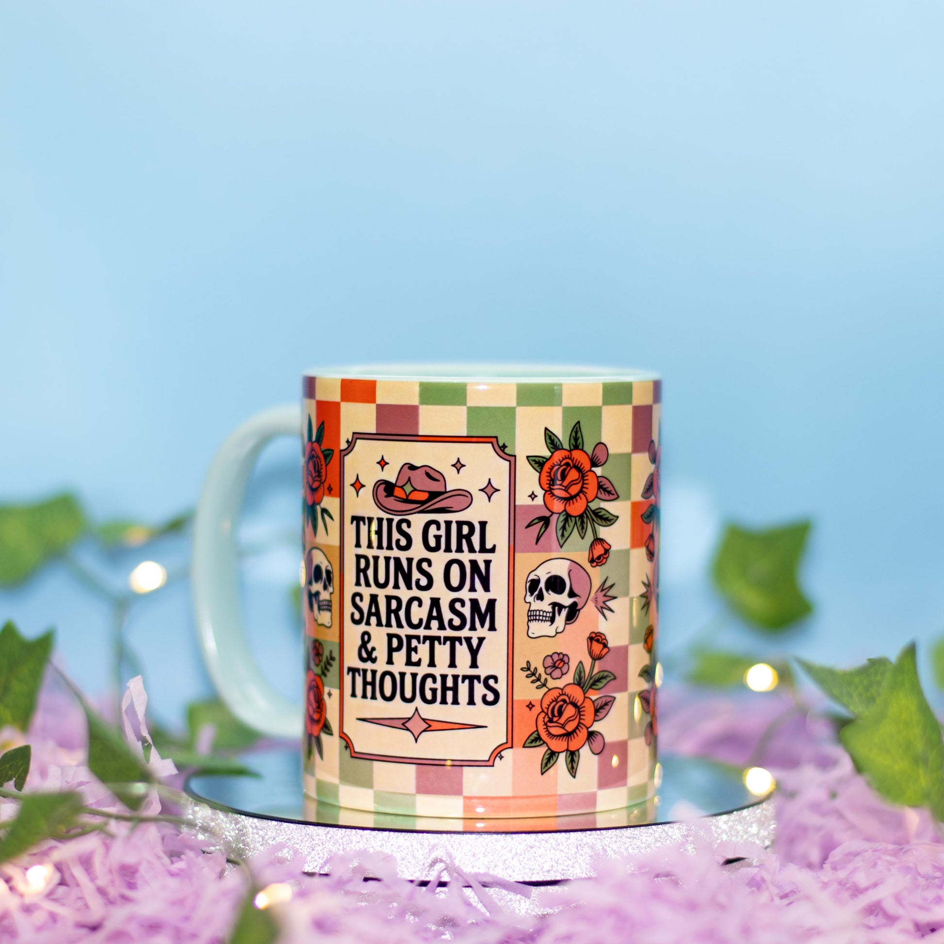 A ceramic mug with a colorful checkered border and illustrations of a cowboy hat, skull, and roses, featuring the text 'THIS GIRL RUNS ON SARCASM & PETTY THOUGHTS'. The mug is styled on a silver tray with purple shredded paper and green leaves against a soft blue background.