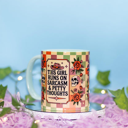 A ceramic mug with a colorful checkered border and illustrations of a cowboy hat, skull, and roses, featuring the text 'THIS GIRL RUNS ON SARCASM & PETTY THOUGHTS'. The mug is styled on a silver tray with purple shredded paper and green leaves against a soft blue background.
