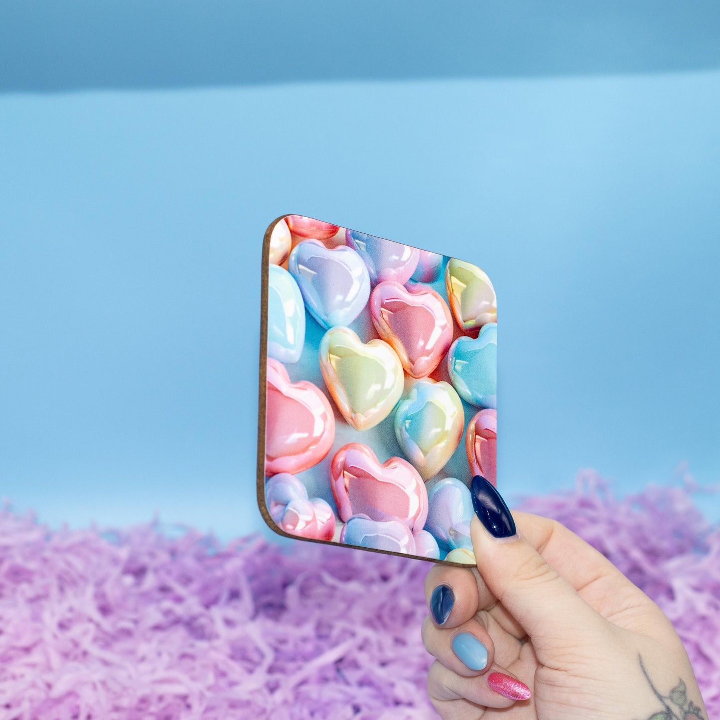Square hard backed coaster featuring a colourful pattern of glossy pastel heart candies, set against a blue background with purple shredded paper.