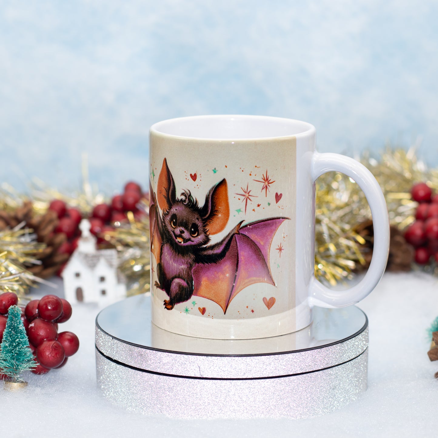 A white ceramic mug featuring a cute bat illustration, displayed on a round silver glitter pedestal 