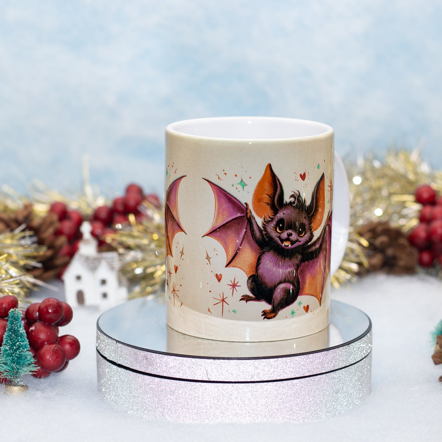 A ceramic mug featuring a detailed illustration of a cute bat with purple wings and orange ears, displayed on a round silver glitter stand