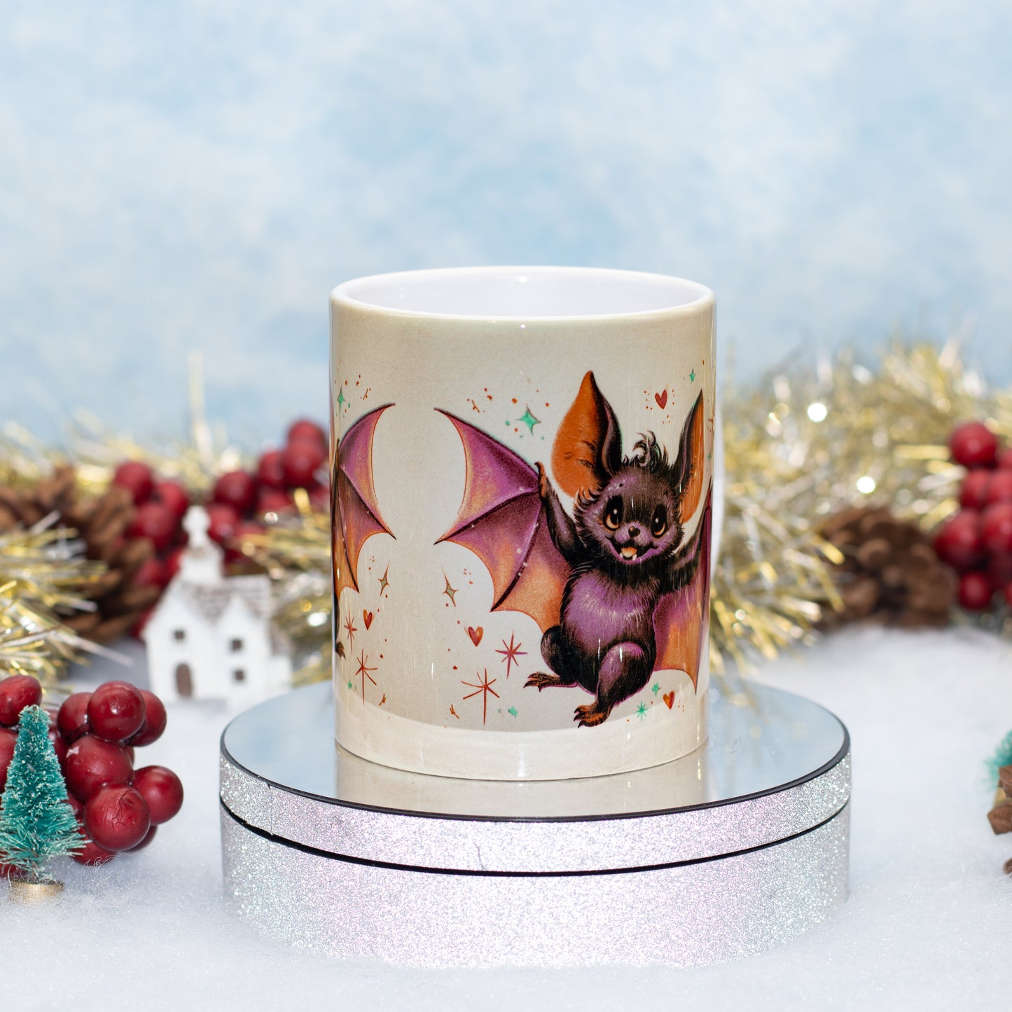 A ceramic mug featuring a cute bat illustration, displayed on a silver glittery round stand 