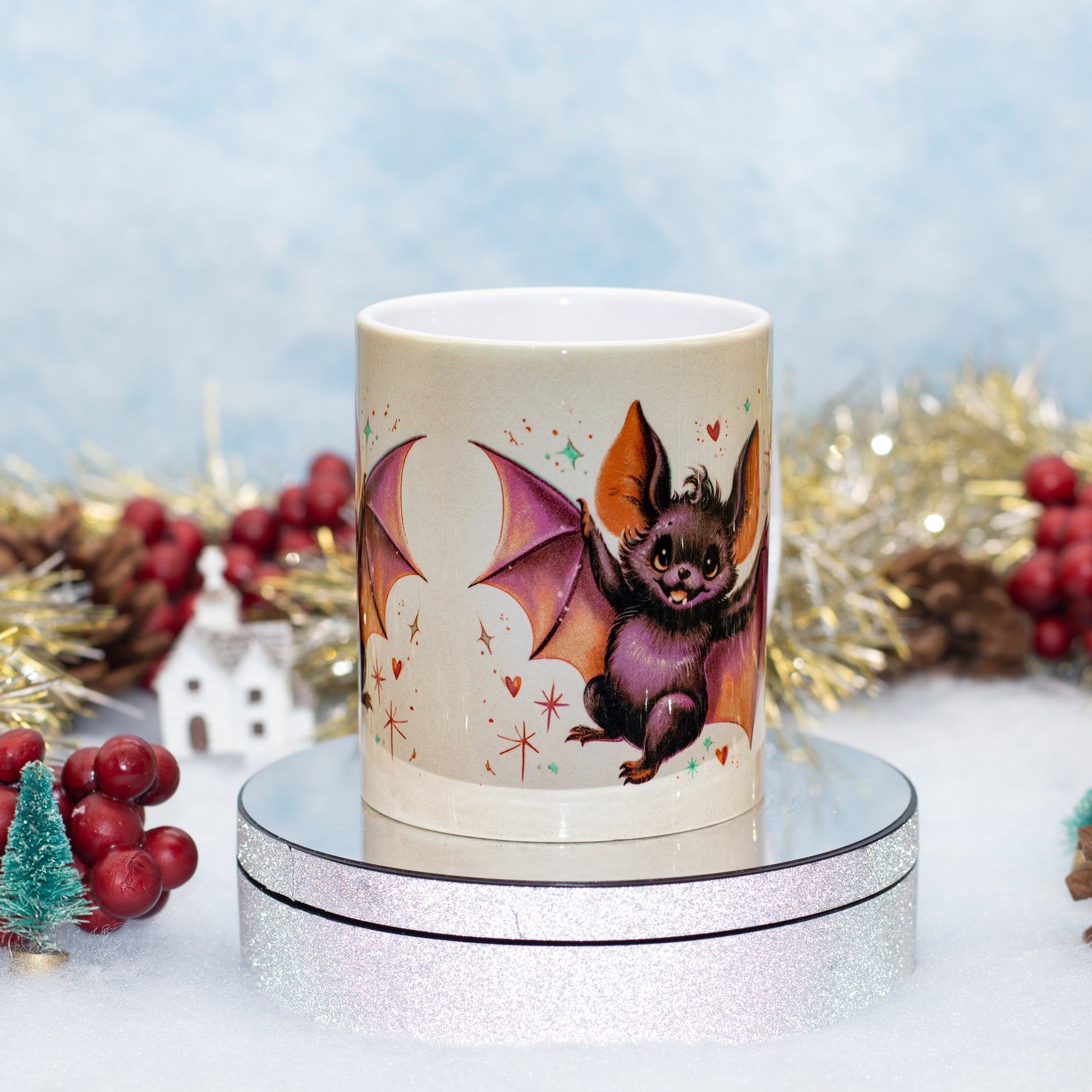 A ceramic mug featuring a cute bat illustration, displayed on a silver glittery round stand 