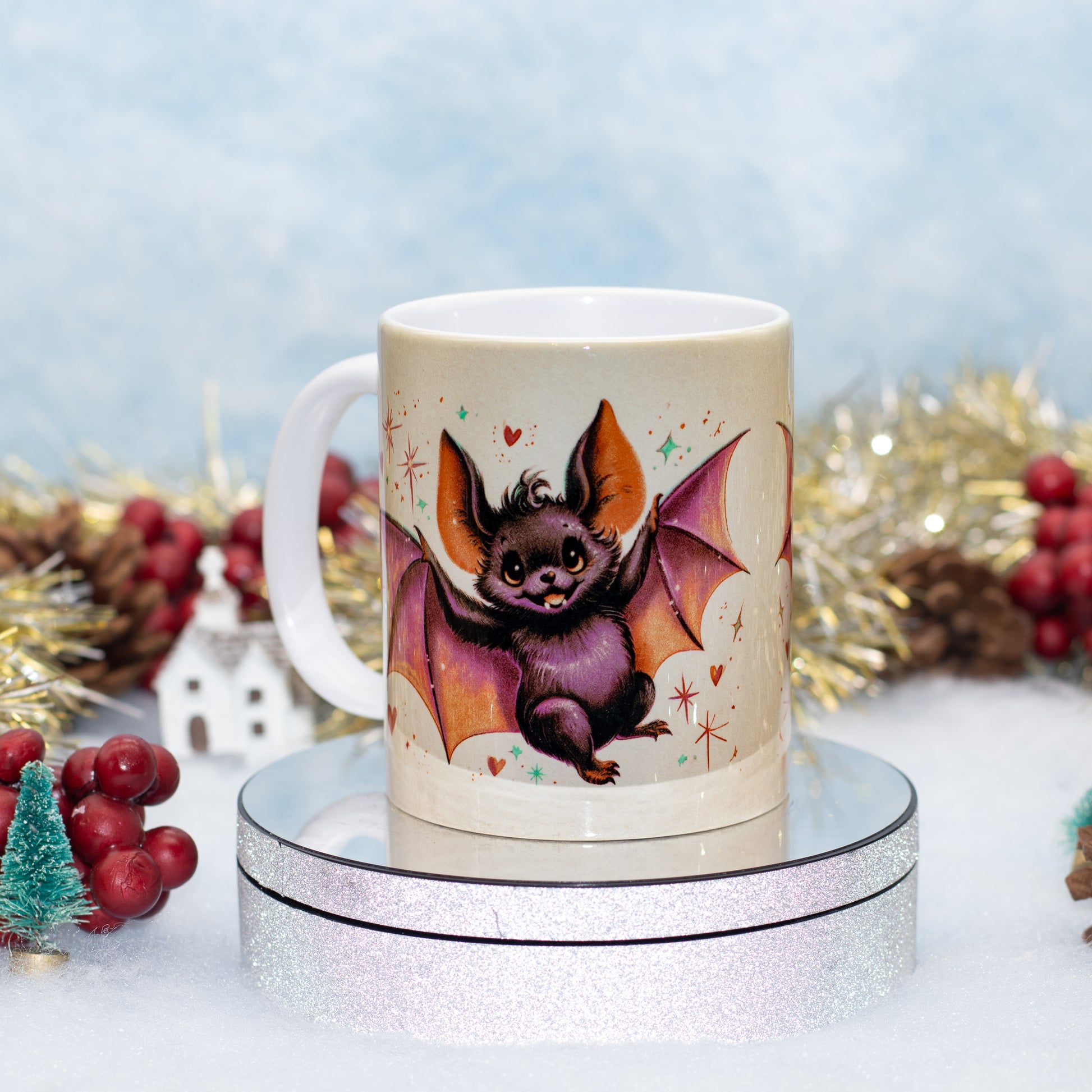 A white ceramic mug featuring a whimsical illustration of a cute purple bat with orange wings, displayed on a round silver glitter pedestal