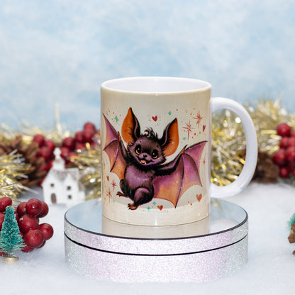 A white ceramic mug featuring a cute cartoon bat illustration, displayed on a round silver glitter pedestal with festive holiday decorations in the background.