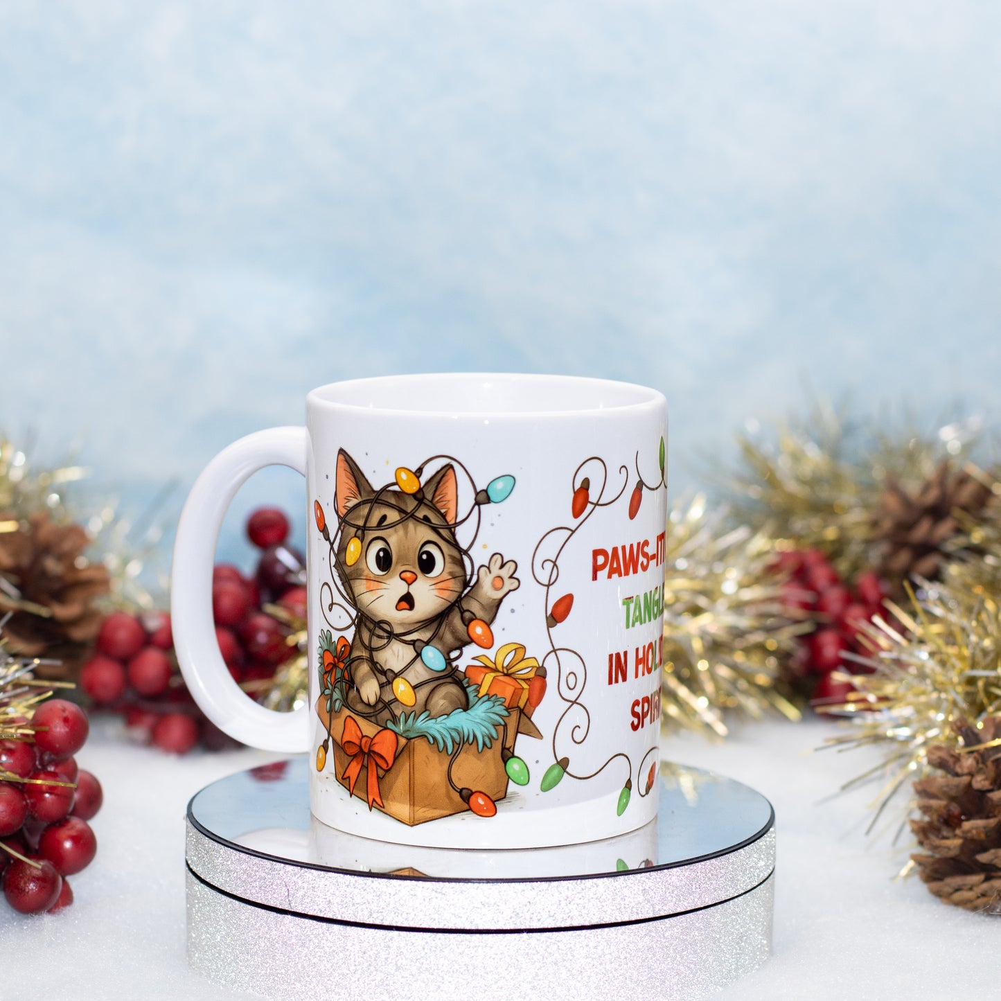 A white ceramic mug featuring a cute cartoon cat surrounded by Christmas gifts and holiday lights, displayed on a round silver stand with festive tinsel and red berries in the background.