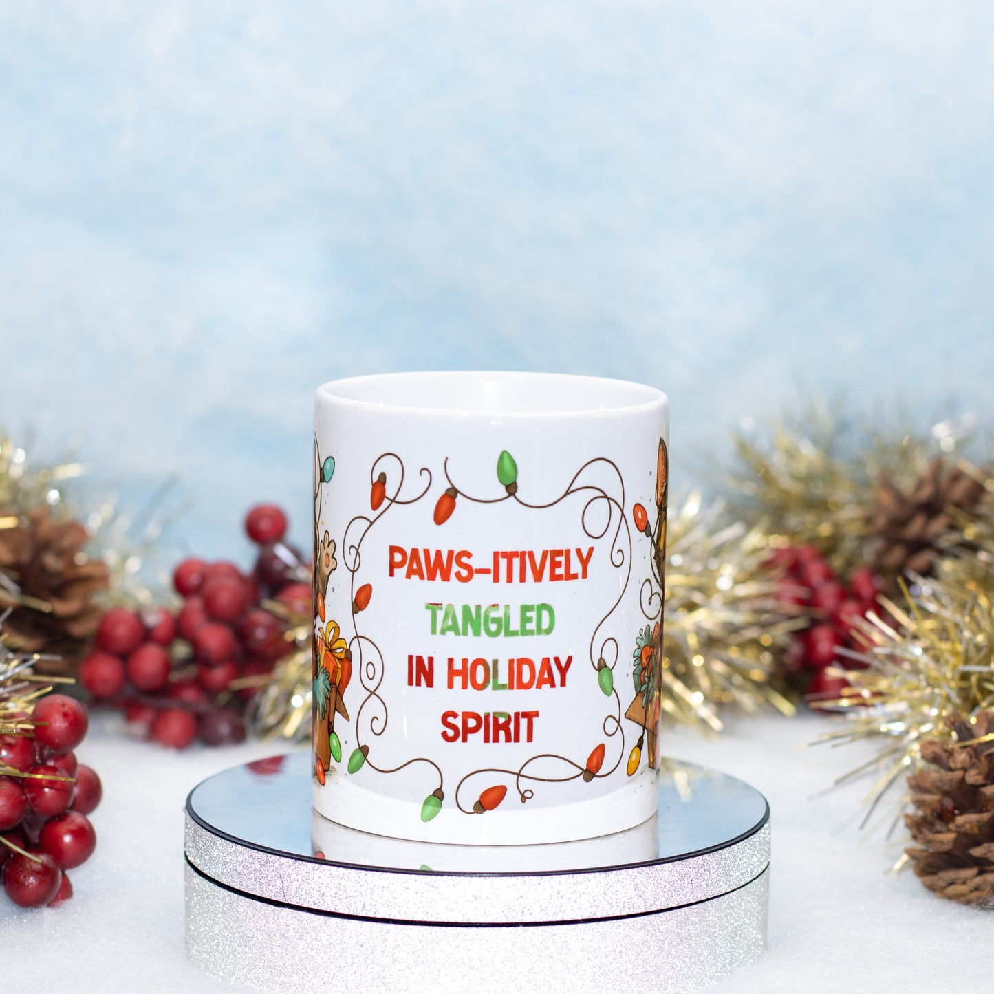 A white ceramic mug with a holiday design and the text 'PAWS-ITIVELY TANGLED IN HOLIDAY SPIRIT' in red and green lettering, displayed on a silver round stand with festive tinsel, pinecones, and red berries in the background.