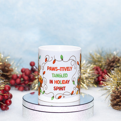 A white ceramic mug with a holiday design and the text 'PAWS-ITIVELY TANGLED IN HOLIDAY SPIRIT' in red and green lettering, displayed on a silver round stand with festive tinsel, pinecones, and red berries in the background.