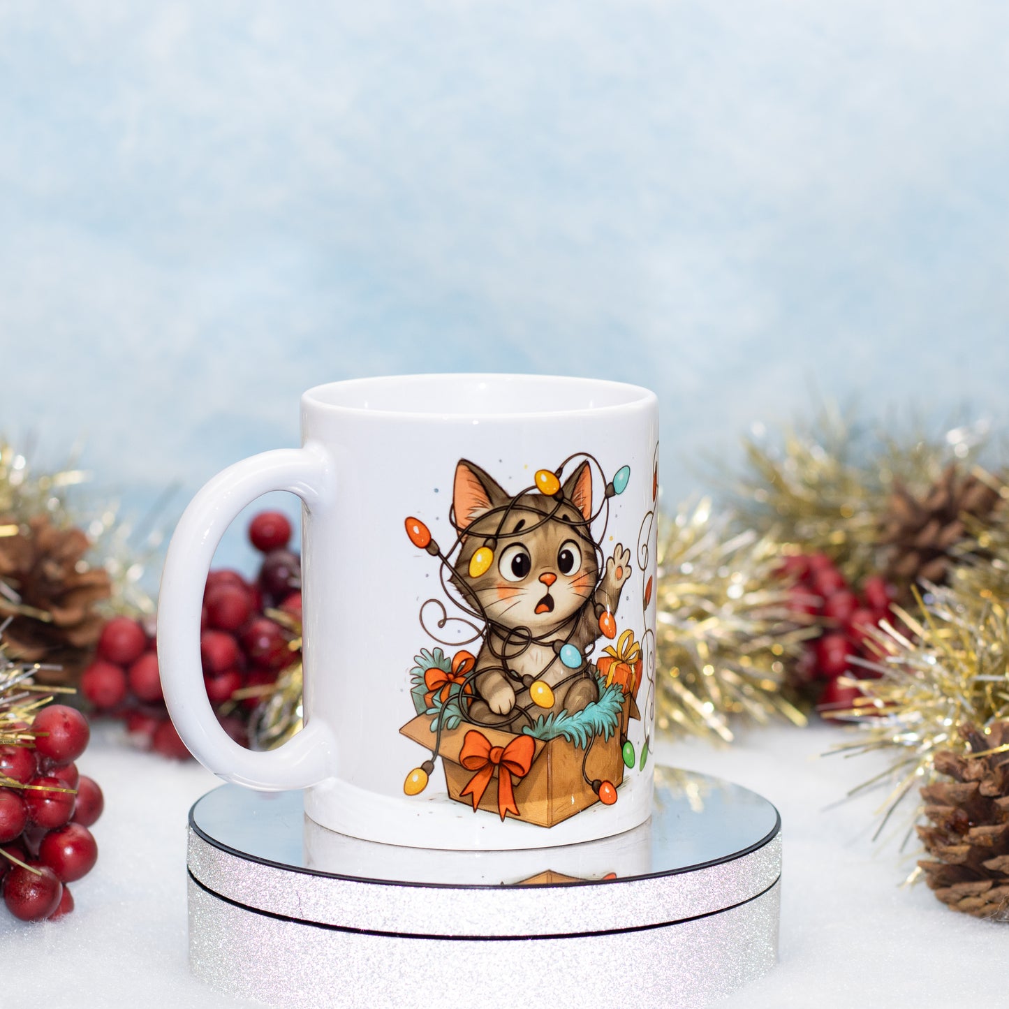 A white ceramic mug featuring a cute cartoon cat illustration sitting in a gift box with colorful holiday lights, displayed on a round silver pedestal with festive Christmas decorations in the background.