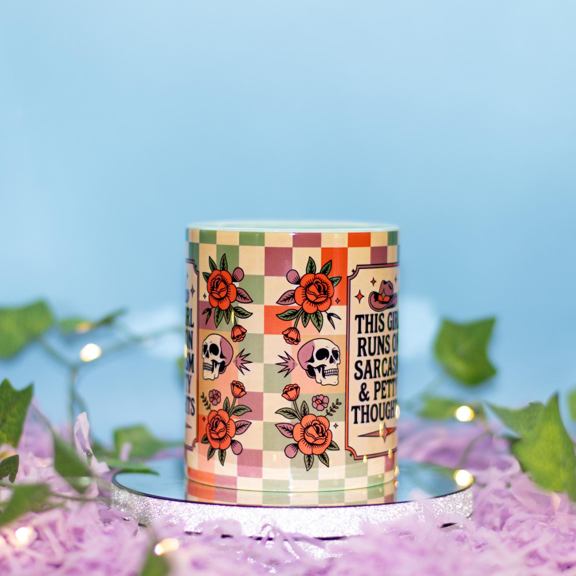 A cylindrical candle with a colorful retro checkered pattern, orange roses, skulls, and the text 'THIS GIRL RUNS ON SARCASM & PETTY THOUGHTS' sits on a silver mirrored base surrounded by purple shredded paper and green leaves against a light blue background.