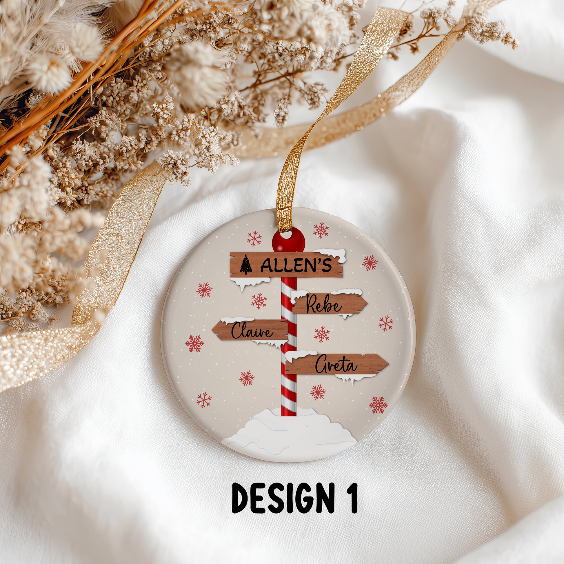 A round personalized Christmas ornament featuring a festive signpost with names on wooden signs, decorated with red snowflakes on a cream background. The ornament hangs from a gold ribbon and is displayed on a white fabric background with dried flowers and a gold ribbon.