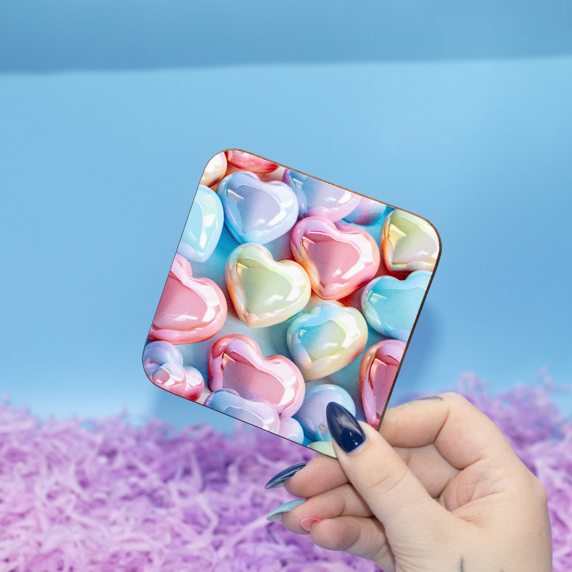 Square hard backed coaster featuring a colourful pattern of glossy pastel heart candies, set against a blue background with purple shredded paper.