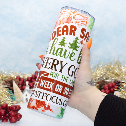 A hand holds a tall insulated tumbler with a white background and colorful Christmas-themed text and illustrations. The tumbler reads 'DEAR SANTA I have been VERY GOOD FOR THE LAS WEEK OR SO LONG JUST FOCUS ON THAT