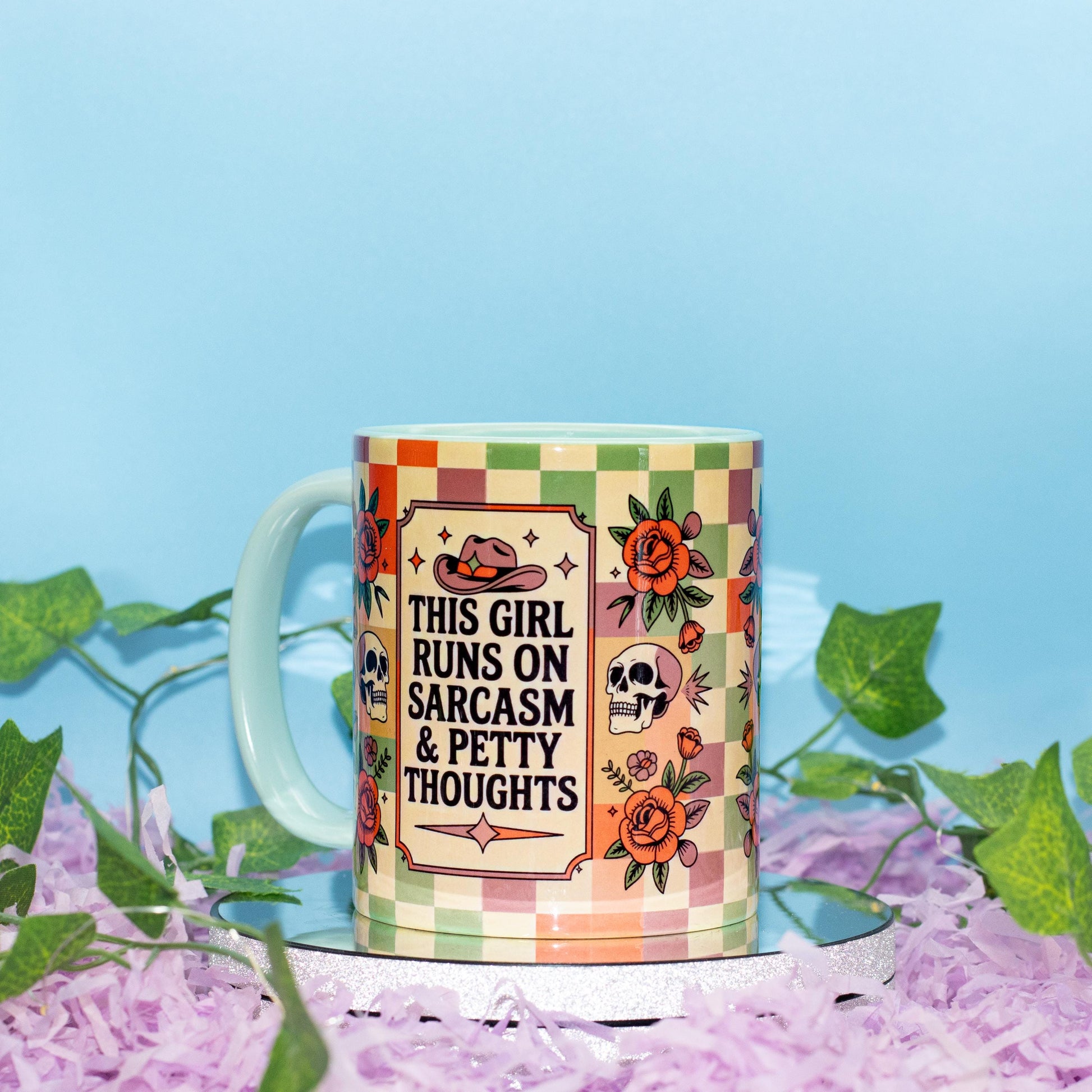A ceramic mug with a colorful checkered pattern and illustrations of a cowboy hat, roses, and skulls, featuring the text 'THIS GIRL RUNS ON SARCASM & PETTY THOUGHTS'. The mug is styled on a white surface covered in purple shredded paper, with green ivy leaves against a light blue background.