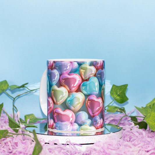 A ceramic mug with a colorful print of glossy, pastel heart-shaped candies, displayed on a mirrored stand surrounded by pink shredded paper and green ivy against a light blue background.