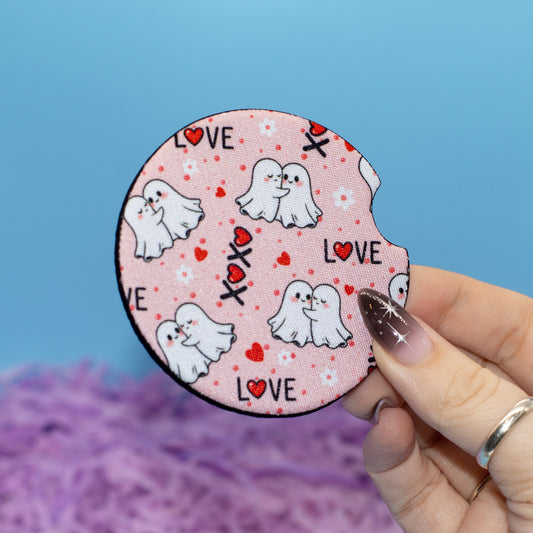 A hand holds a round coaster featuring a cute cartoon ghost pattern, hearts, and the word 'LOVE' against a blue background with purple fluffy decoration.