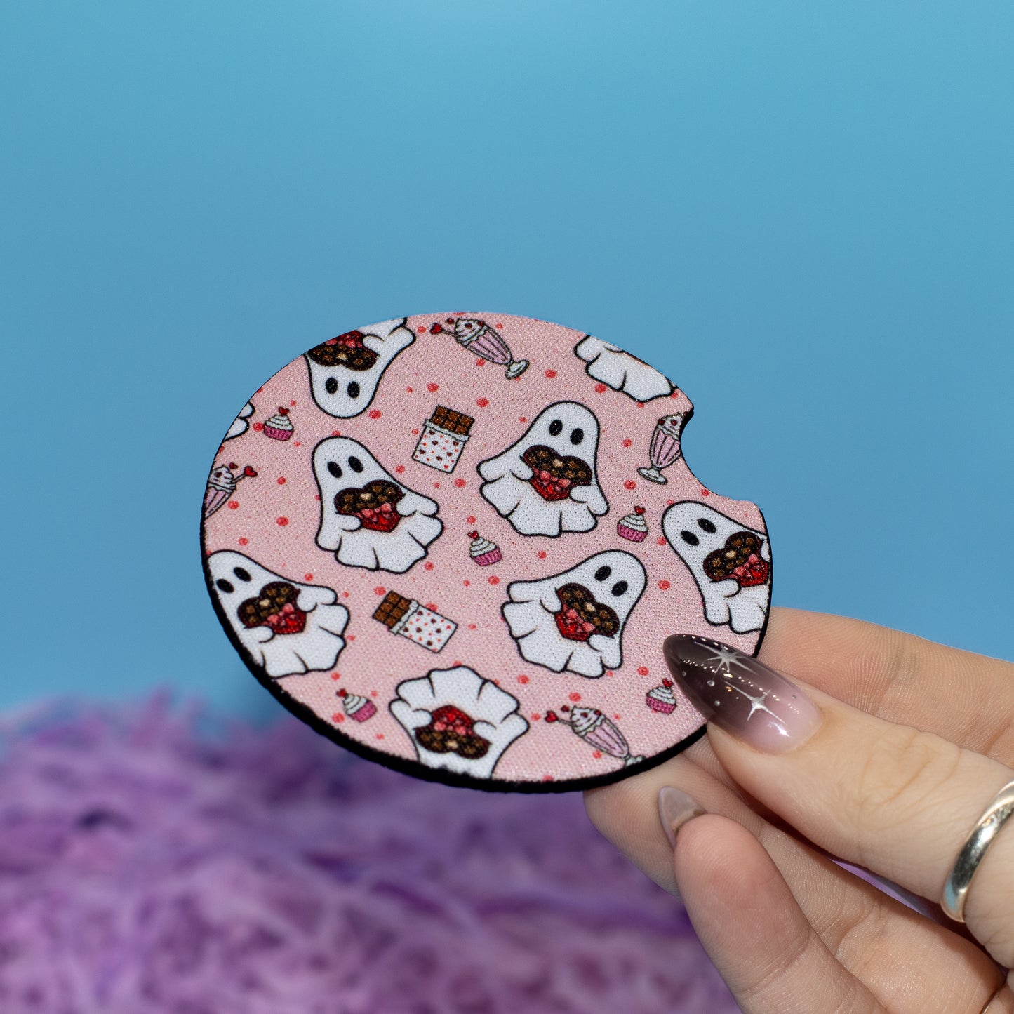 A hand holds a round car coaster featuring a cute ghost and candy pattern, set against a blue background with purple fluffy decoration.
