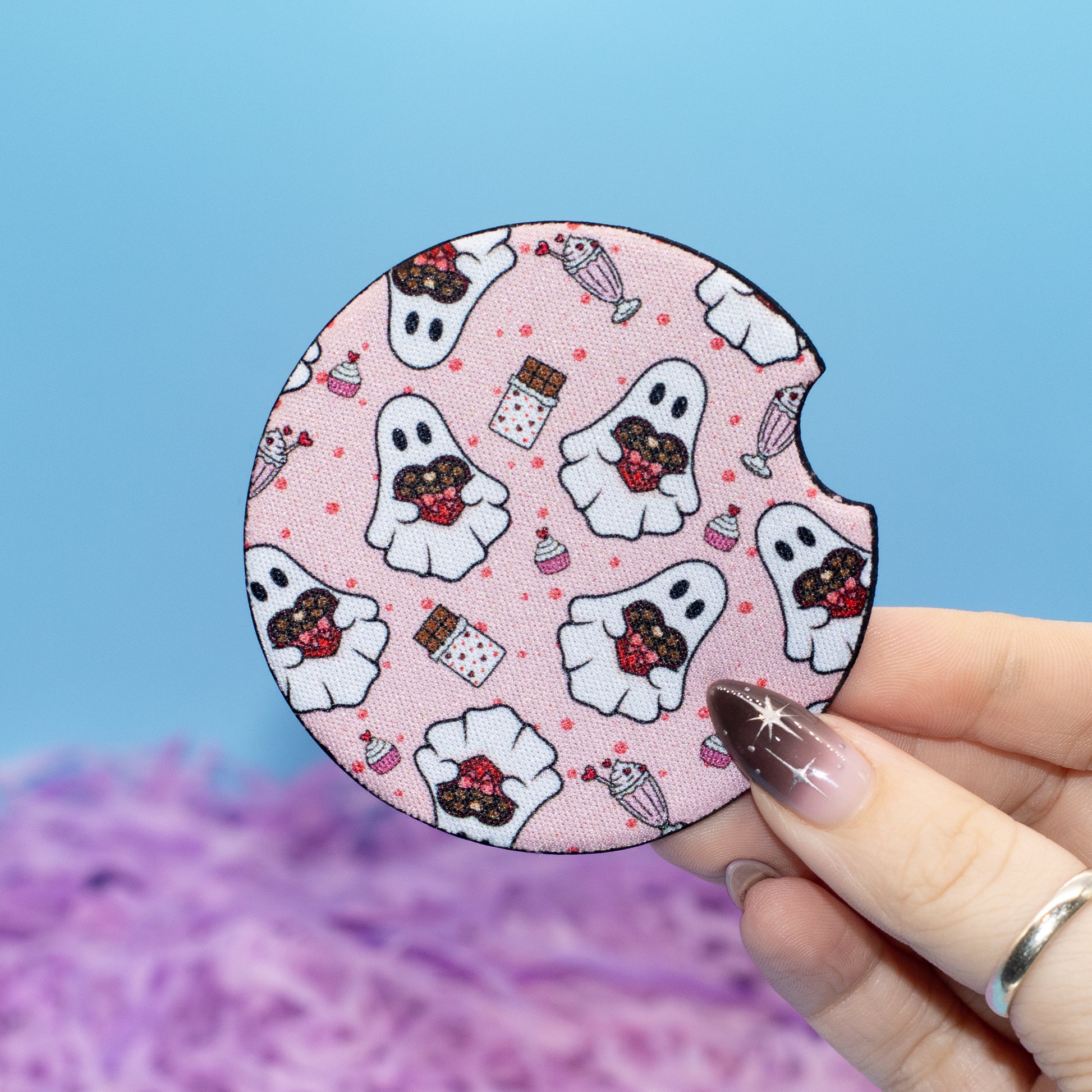A hand holds a round coaster featuring a cute ghost and candy pattern, set against a blue background with purple shredded paper.