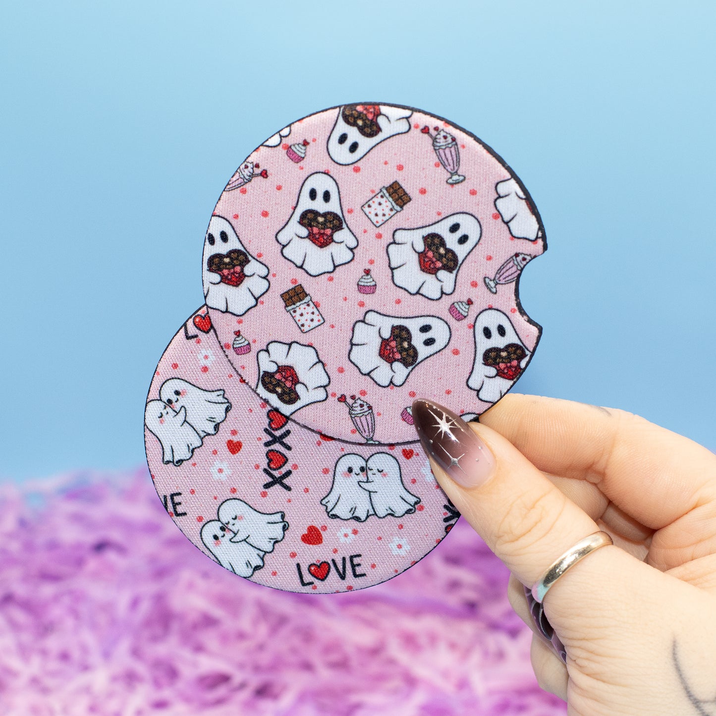 A hand  holds two round coasters featuring a pink and white ghost and candy pattern against a blue and purple background.
