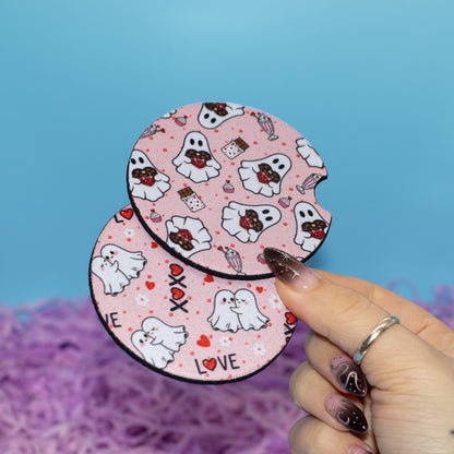 A hand holds two round pink coasters featuring a cute cartoon ghost pattern with small red hearts, chocolate bars, and the word 'LOVE' against a blue background with purple shredded paper.