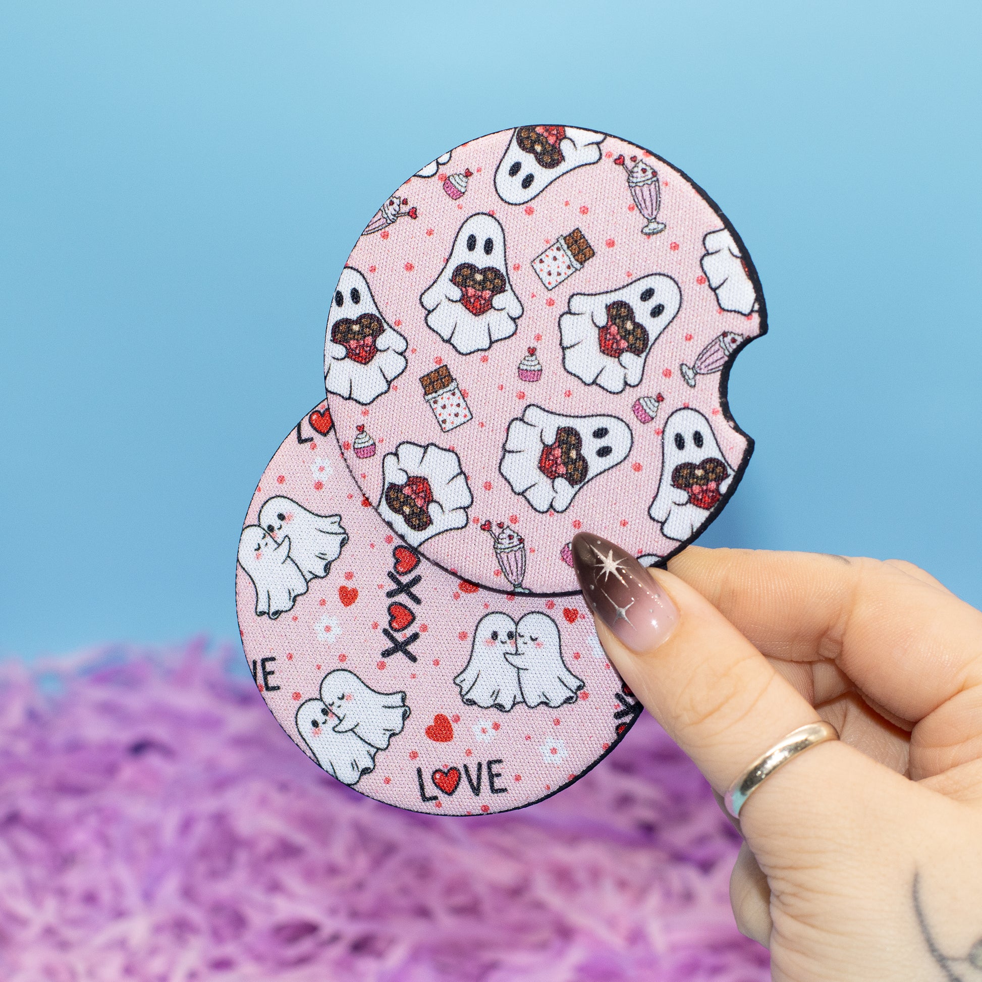 A hand holds two round, car coaster with a pink Halloween-themed pattern featuring cartoon ghosts, hearts, and candy. The background is a solid light blue with purple shredded paper at the bottom.