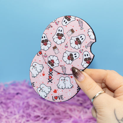 A hand holds two round, car coaster with a pink Halloween-themed pattern featuring cartoon ghosts, hearts, and candy. The background is a solid light blue with purple shredded paper at the bottom.