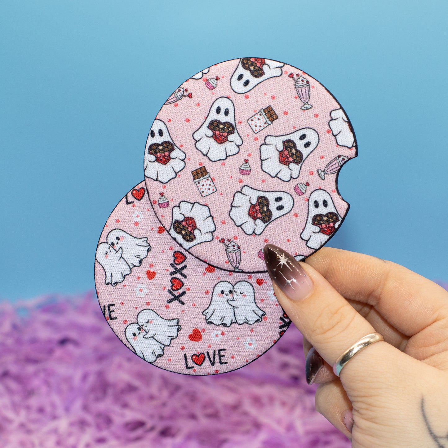 A hand holds two round, pink Halloween-themed coasters featuring cartoon ghosts, chocolate-covered treats, and hearts against a blue background with purple shredded paper.