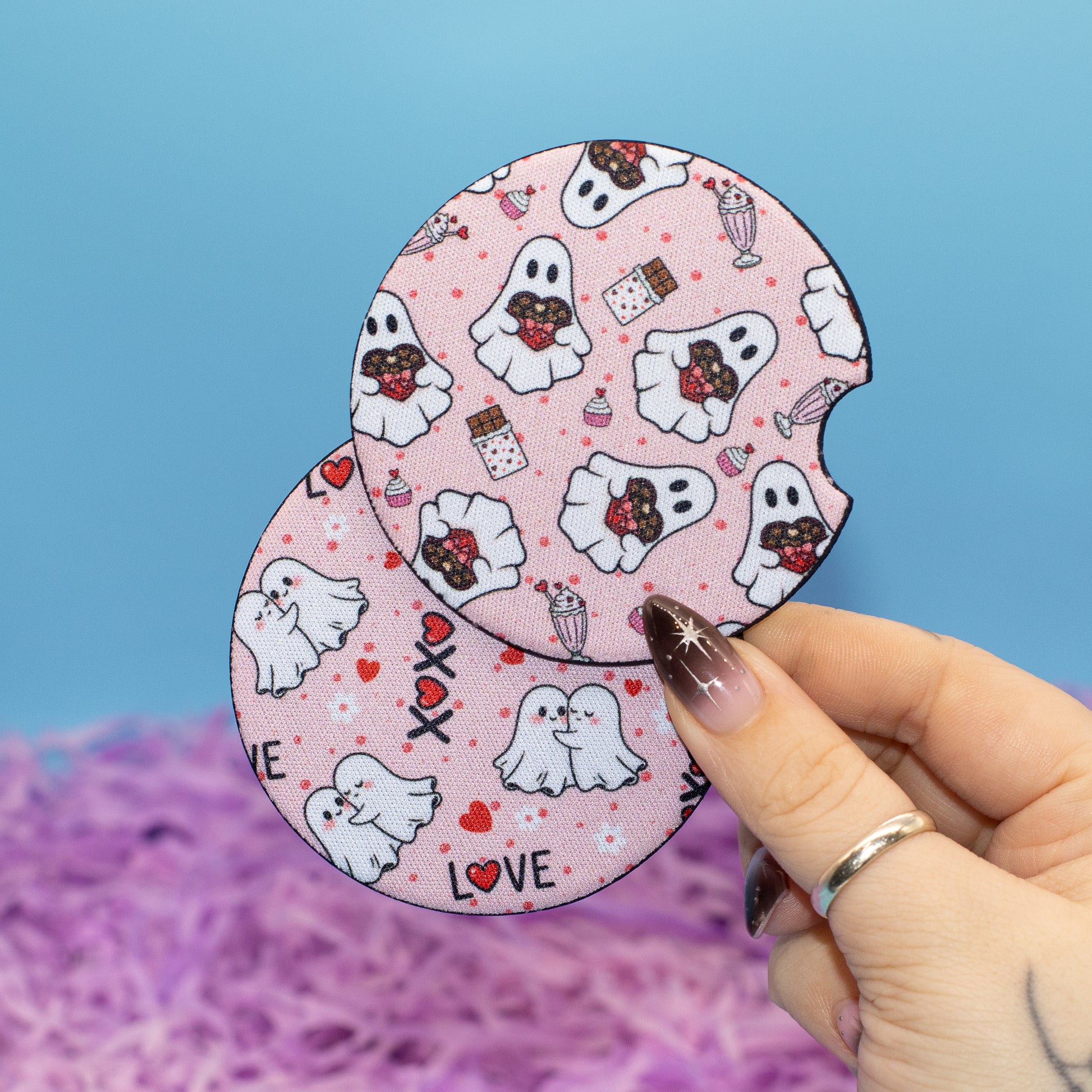 A hand holds two round, pink Halloween-themed coasters featuring cartoon ghosts, chocolate-covered treats, and hearts against a blue background with purple shredded paper.