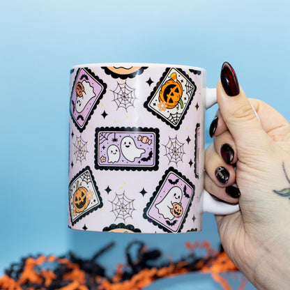 a white ceramic mug decorated with a Halloween pattern of purple ghosts, orange pumpkins, and black spiderwebs against a light blue background.