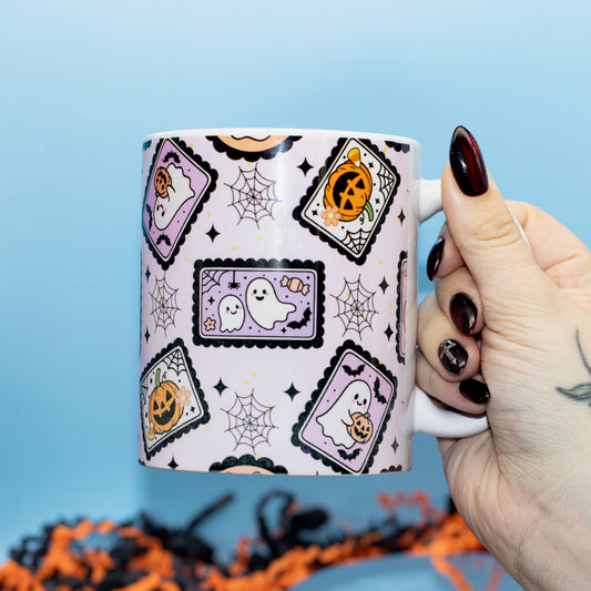 a white ceramic mug decorated with a Halloween pattern of purple ghosts, orange pumpkins, and black spiderwebs against a light blue background.