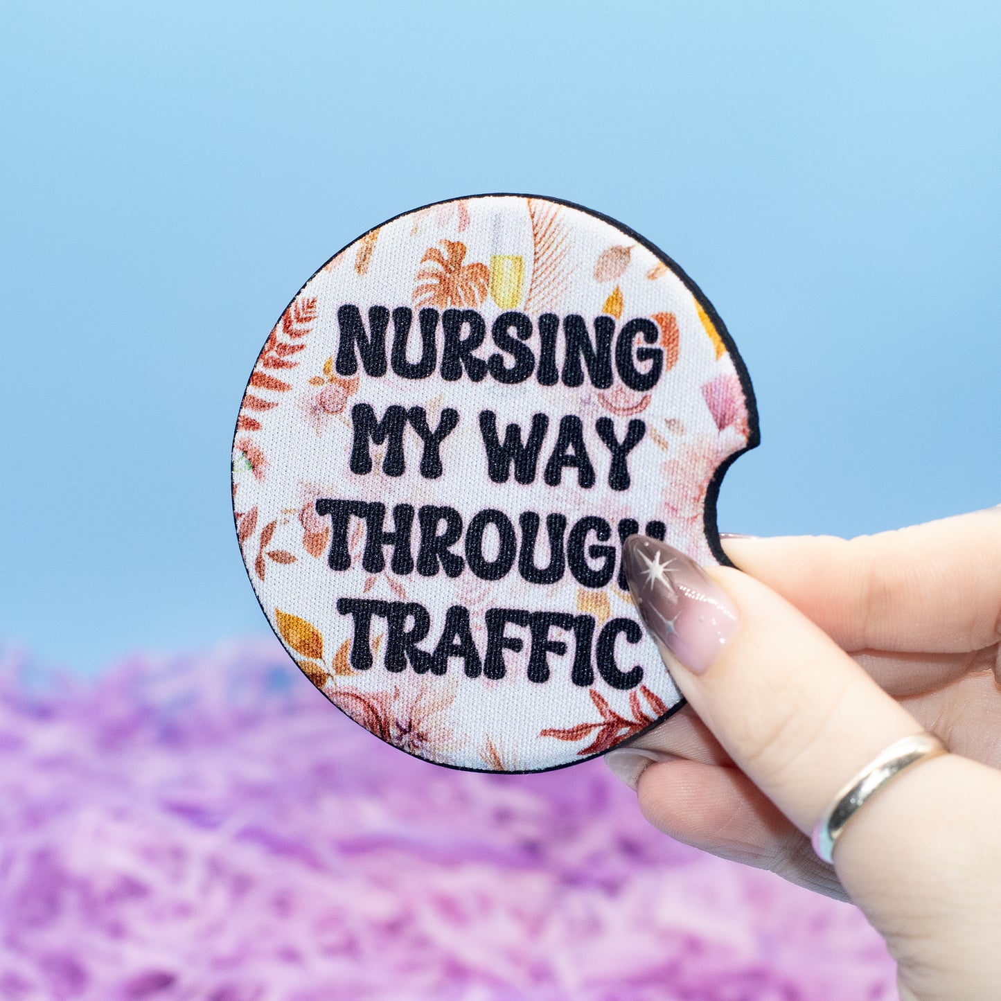 A hand holds a round car coaster with the text 'NURSING MY WAY THROUGH TRAFFIC' in black letters over a floral patterned background. The hand is in front of a blurred blue and purple background.
