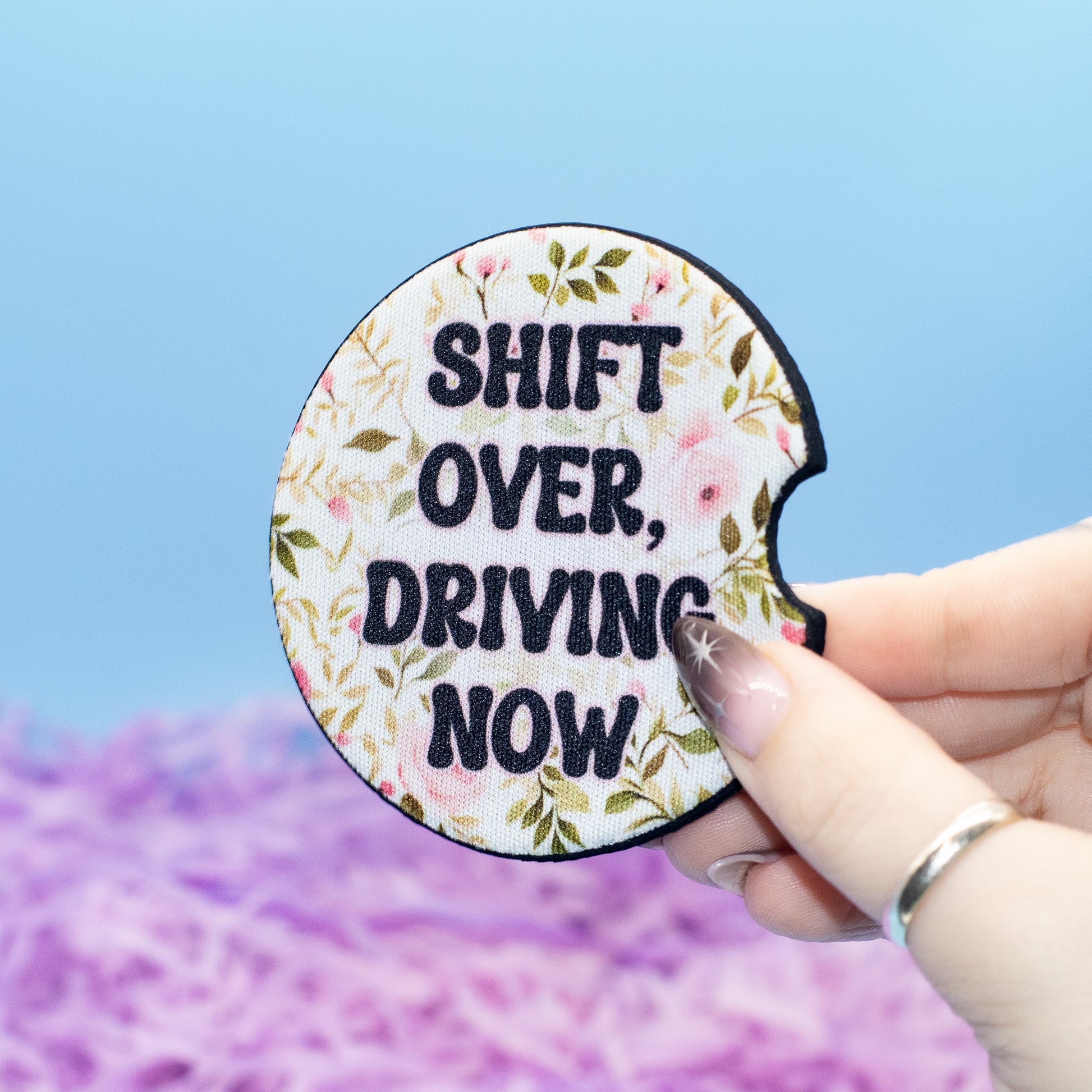 A hand holds a round car coaster with the text 'SHIFT OVER, DRIVING NOW' in black lettering over a floral cream background. 