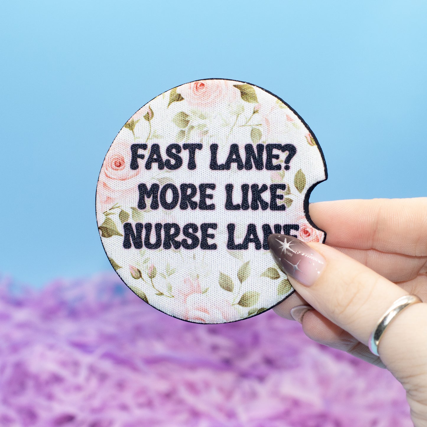 A hand holds a round embroidered patch with a floral design and the text 'FAST LANE? MORE LIKE NURSE LANE!' against a blue and purple background.