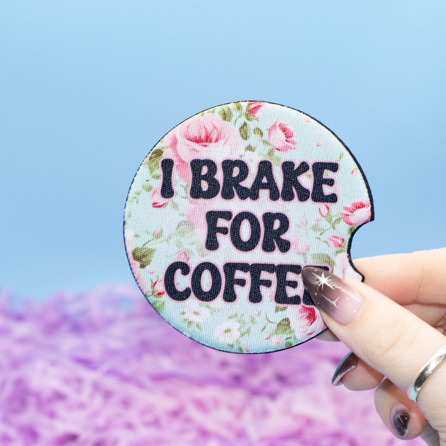 A hand holds a round car coaster  that reads 'I BRAKE FOR COFFEE' in black letters over a pink and green floral pattern on a light blue background. The background is a soft-focus blue and purple scene.