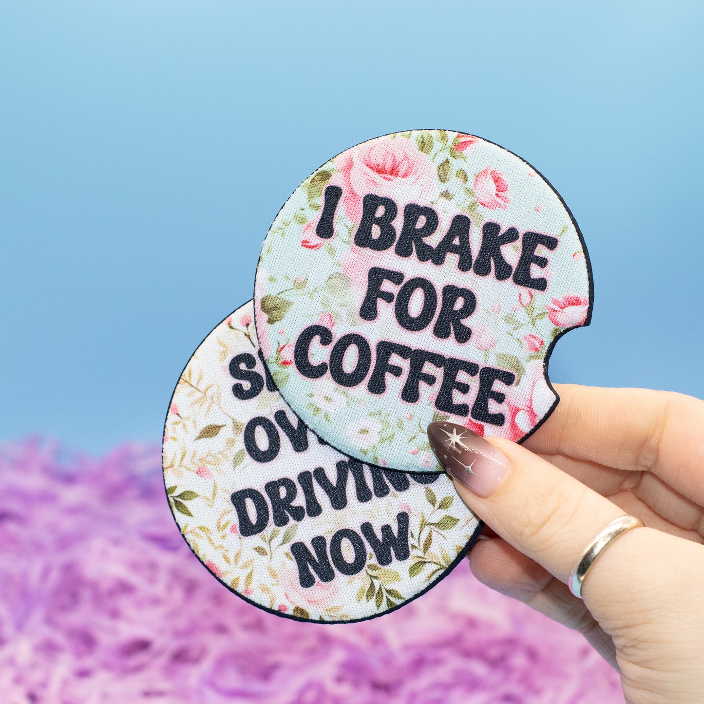 Two round car coaster with a floral print and the text 'I BRAKE FOR COFFEE' and 'SHIFT OVER DRIVING NOW', held by a hand against a blue sky and purple flower background.