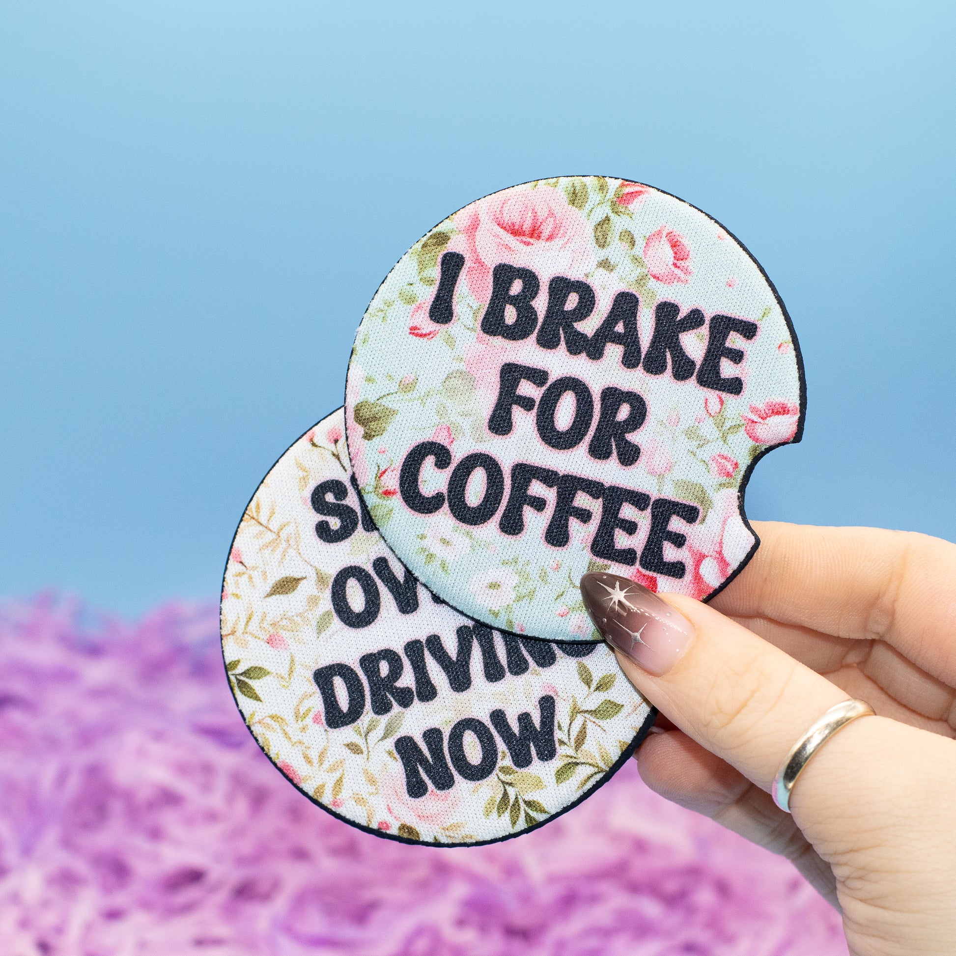 Two round car coaster with a floral print and the text 'I BRAKE FOR COFFEE' and 'SHIFT OVER DRIVING NOW', held by a hand against a blue sky and purple flower background.