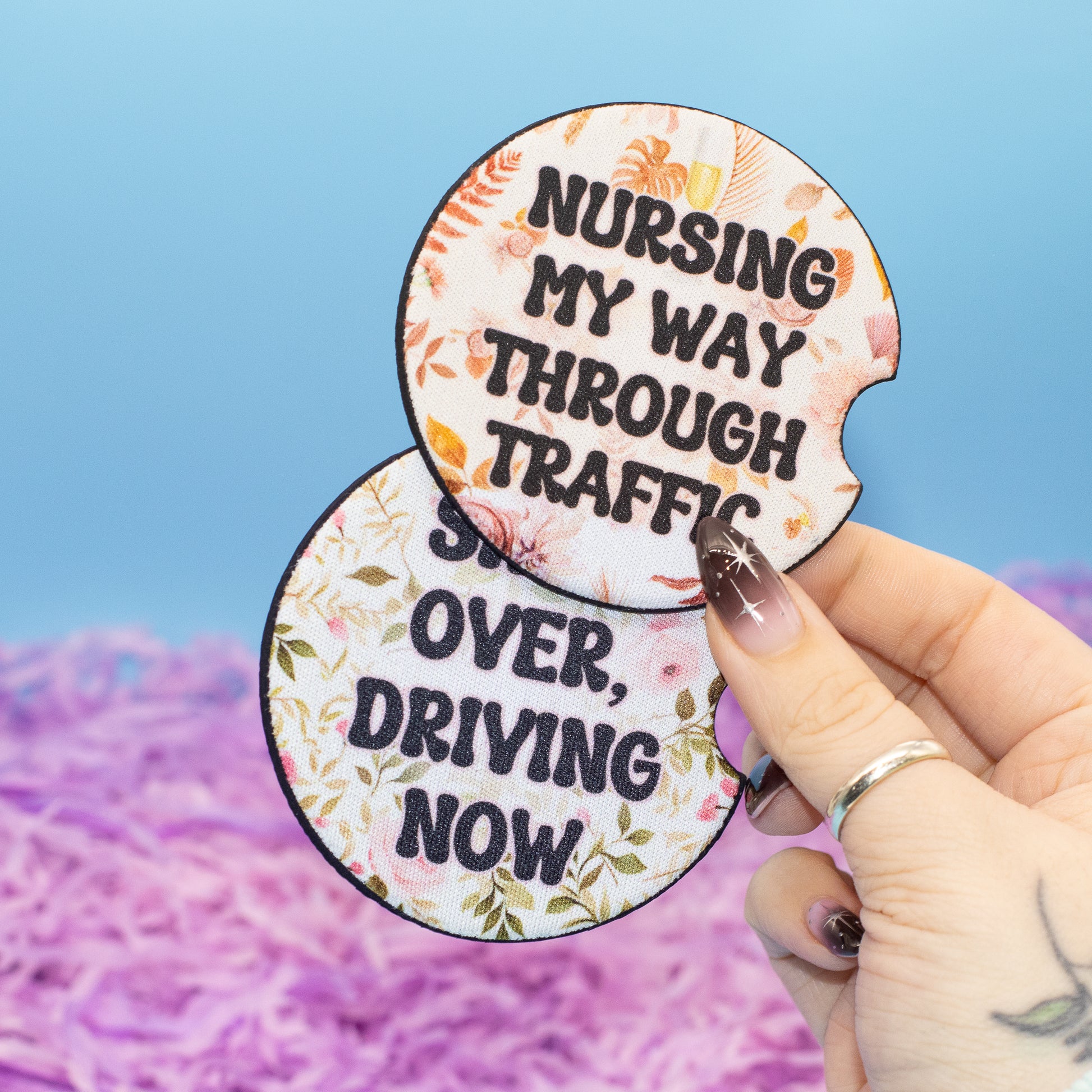 A hand holds two round car coasters with floral borders and humorous nursing-themed text against a blue and pink background.