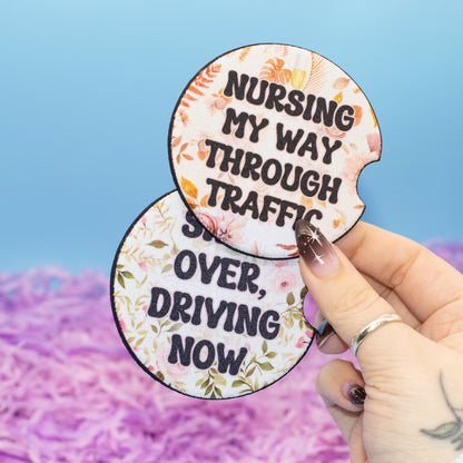A hand holds two round car coasters with floral borders and humorous nursing-themed text against a blue and pink background.