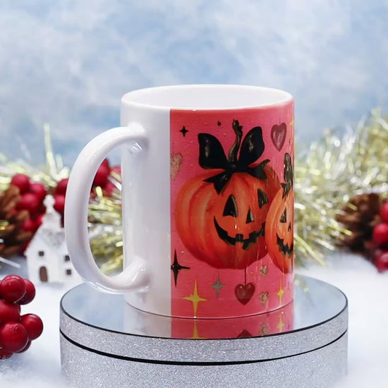 Video showing A white ceramic mug with a pink Halloween-themed print featuring jack-o'-lantern pumpkins, hearts, and stars