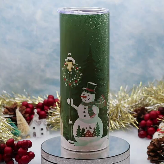 Video Showcasing A hand holds a green insulated tumbler decorated with a winter scene featuring a snowman, Christmas wreath, street lamp, evergreen trees, and small houses